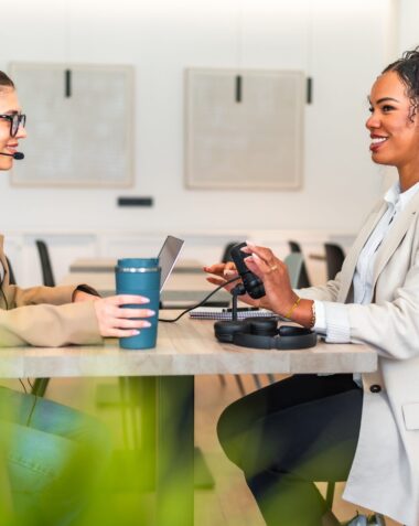 Two podcasters are recording a new episode in a modern office, using a professional microphone and laptop
